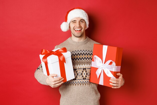 Concepto de vacaciones de invierno, año nuevo y celebración. Retrato de joven confiado y descarado preparado regalos para Navidad, guiñando un ojo y sosteniendo regalos, de pie sobre fondo rojo.