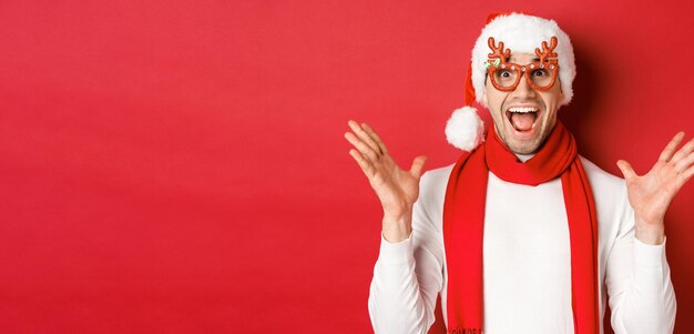 Concepto de vacaciones de invierno y celebración navideña Imagen de un hombre sorprendido y feliz que se ve asombrado con gafas de fiesta y disfrutando el año nuevo de pie sobre un fondo rojo