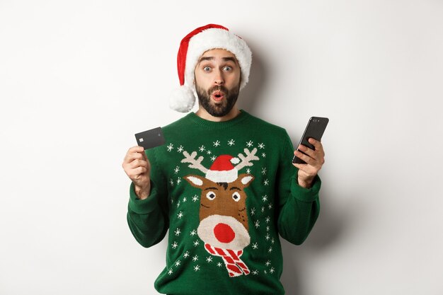 Concepto de vacaciones de invierno y compras en línea. Hombre sorprendido comprando regalos en internet con teléfono móvil y tarjeta de crédito, de pie en el sombrero de Santa sobre fondo blanco.