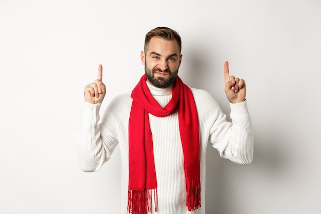 Concepto de vacaciones de invierno y compras de Navidad. Chico disgustado y escéptico quejándose, señalando con el dedo a un producto malo, de pie contra el fondo blanco.