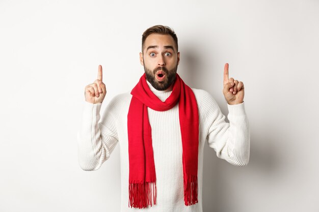 Concepto de vacaciones de invierno y compras de Navidad. Modelo masculino sorprendido con barba apuntando con el dedo hacia arriba, diciendo wow, mirando impresionado a la cámara, fondo blanco.