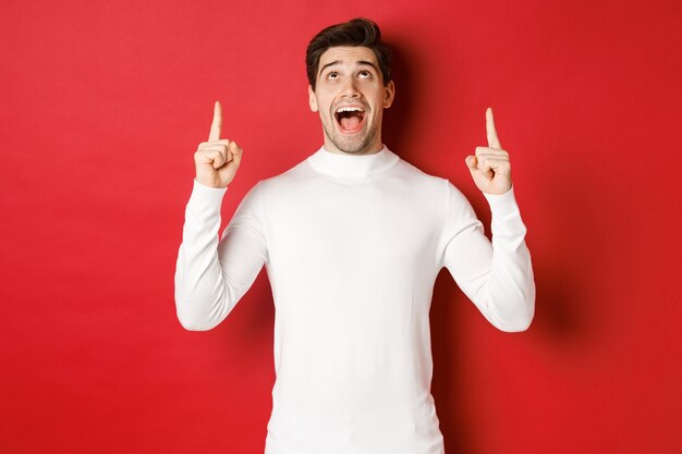 Concepto de vacaciones de invierno. Imagen de un chico guapo sorprendido reaccionando al anuncio de Navidad, apuntando y mirando hacia el espacio de la copia, de pie sobre fondo rojo.