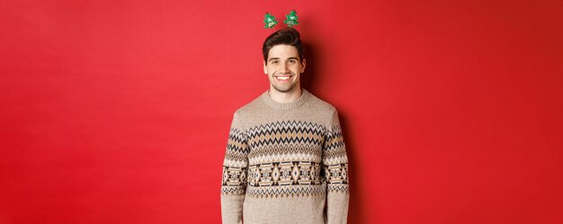 Concepto de vacaciones de invierno, navidad y celebración. Guapo chico barbudo en suéter deseando feliz año nuevo, sonriendo a la cámara, de pie sobre fondo rojo.