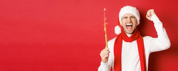 Concepto de vacaciones de invierno de navidad y celebración Hombre guapo celebrando el año nuevo y divirtiéndose sosteniendo bengala y sonriendo con sombrero de santa de pie sobre fondo rojo