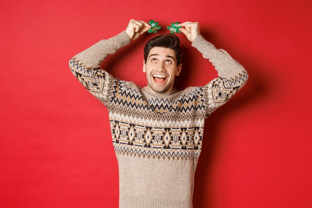 Concepto de vacaciones de invierno, navidad y celebración. Imagen de hombre adulto divertido y lindo disfrutando de la fiesta de año nuevo, sonriendo y regocijándose, de pie sobre fondo rojo.