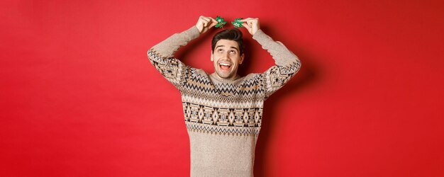 Concepto de vacaciones de invierno, navidad y celebración. Imagen de un hombre adulto divertido y lindo disfrutando de la fiesta de año nuevo, sonriendo y regocijándose, de pie sobre un fondo rojo.