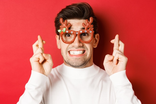 Concepto de vacaciones de invierno, navidad y celebración. Primer plano de hombre esperanzado en gafas de fiesta, cruzando los dedos y pidiendo un deseo en año nuevo, de pie sobre fondo rojo.