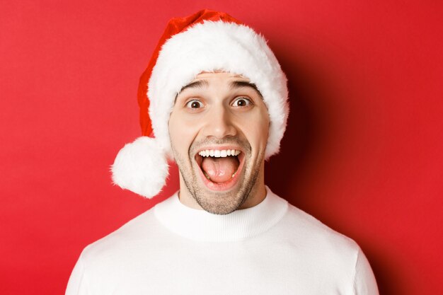 Concepto de vacaciones de invierno, navidad y celebración. Primer plano de un hombre guapo sorprendido con sombrero de santa, escuchar la increíble oferta de promoción de año nuevo, de pie contra el fondo rojo.