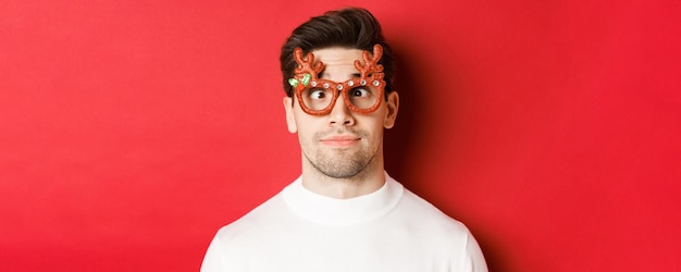 Concepto de vacaciones de invierno, navidad y celebración. Primer plano de morena divertida en gafas de fiesta, entrecerrar los ojos y hacer muecas, de pie sobre fondo rojo.