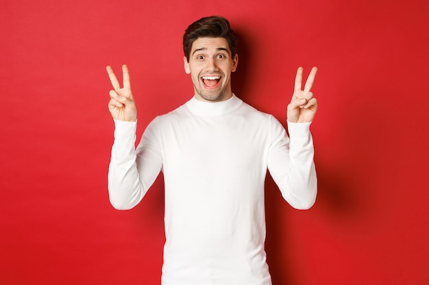 Concepto de vacaciones de invierno navidad y estilo de vida chico divertido guapo en suéter blanco mostrando la paz ...