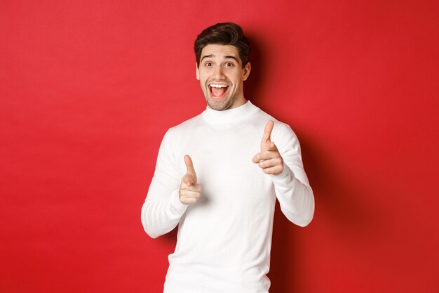 Concepto de vacaciones de invierno, navidad y estilo de vida. Chico guapo alegre en suéter blanco felicitándote, señalando con el dedo a la cámara y deseando feliz año nuevo, de pie sobre fondo rojo