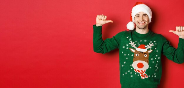 Concepto de vacaciones de invierno Navidad y estilo de vida Feliz chico atractivo con gorro de Papá Noel y suéter apuntándose a sí mismo con cara complacida de pie sobre fondo rojo