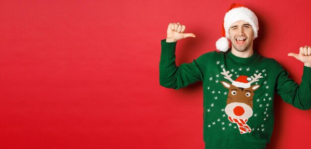 Concepto de vacaciones de invierno, navidad y estilo de vida. Hombre guapo descarado con gorro de Papá Noel y suéter verde, apuntando a sí mismo y guiñando un ojo, de pie sobre fondo rojo.