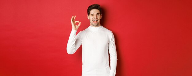 Concepto de vacaciones de invierno, navidad y estilo de vida. Hombre guapo con suéter blanco guiñándote, mostrando un buen signo y sonriendo, garantizando la calidad, recomendando algo