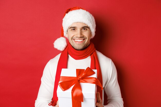 Concepto de vacaciones de invierno, navidad y estilo de vida. Primer plano de atractivo chico barbudo con gorro de Papá Noel y bufanda, sosteniendo un regalo, dando un regalo de año nuevo y sonriendo, fondo rojo