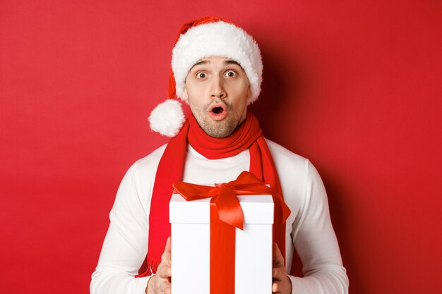 Concepto de vacaciones de invierno, navidad y estilo de vida. Primer plano de chico guapo sorprendido con gorro de Papá Noel y bufanda, mirando sorprendido y sosteniendo un regalo de año nuevo, de pie sobre fondo rojo.