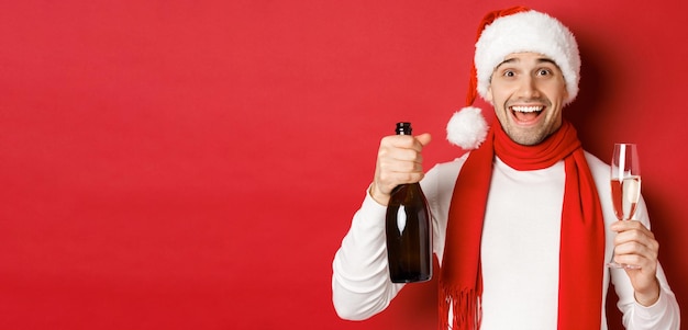 Concepto de vacaciones de invierno navidad y estilo de vida Primer plano de un hombre guapo alegre sosteniendo una botella de champán y una copa celebrando el año nuevo de pie sobre fondo rojo