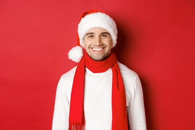 Concepto de vacaciones de invierno, navidad y estilo de vida. Primer plano de un hombre guapo con cerdas, con sombrero de navidad con bufanda y sonriendo feliz, deseando feliz año nuevo, fondo rojo.