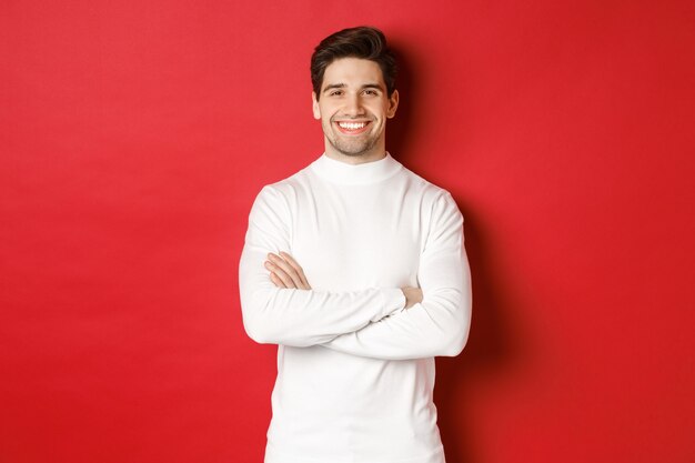 Concepto de vacaciones de invierno, navidad y estilo de vida. Retrato de hombre guapo seguro con cerdas, vestido con suéter blanco, brazos cruzados sobre el pecho y sonriendo satisfecho.