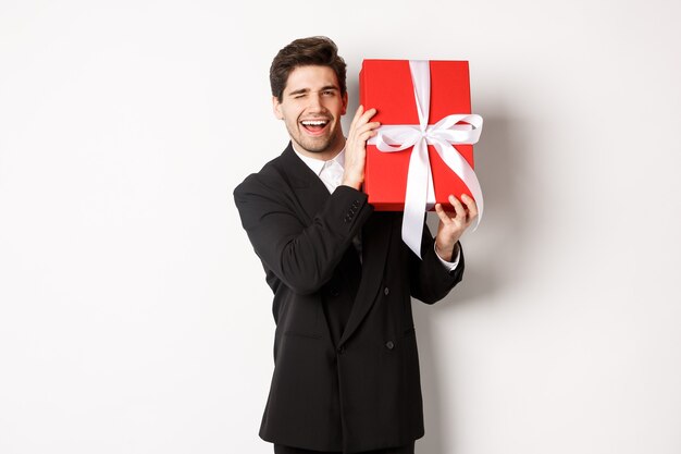 Concepto de vacaciones de Navidad, celebración y estilo de vida. Hombre atractivo en traje negro, sosteniendo un regalo de año nuevo y sonriendo, de pie con un presente sobre fondo blanco.
