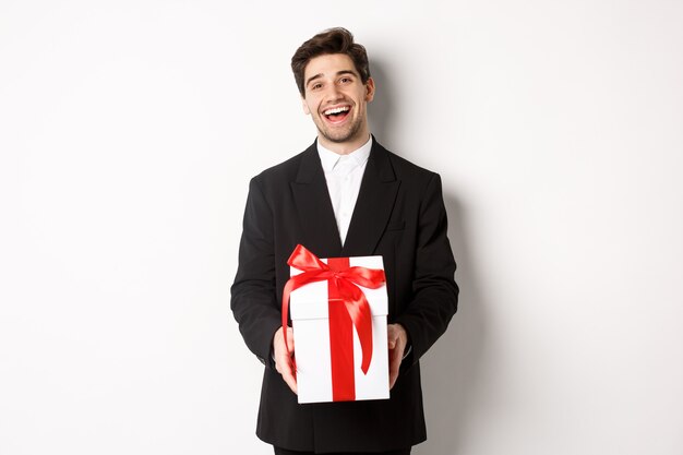 Concepto de vacaciones de Navidad, celebración y estilo de vida. Hombre guapo alegre en traje negro, sosteniendo un regalo de Navidad y sonriendo, de pie contra el fondo blanco.