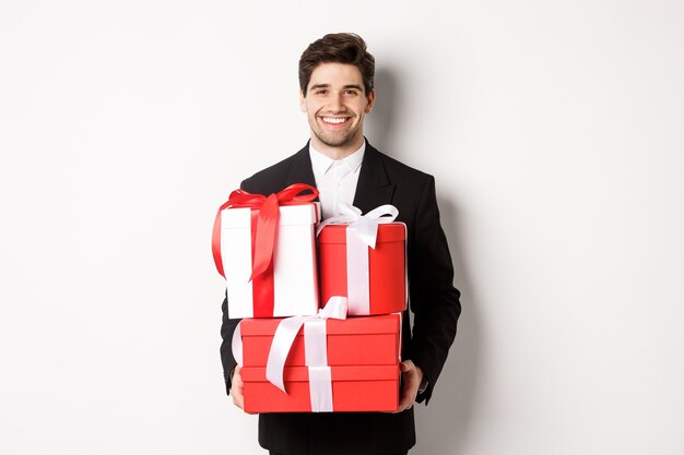 Concepto de vacaciones de Navidad, celebración y estilo de vida. Imagen de atractivo novio en traje negro, sosteniendo regalos y sonriendo, deseando feliz año nuevo, de pie sobre fondo blanco.