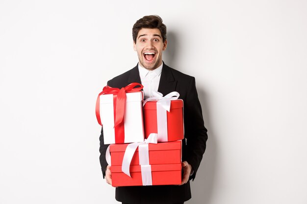 Concepto de vacaciones de Navidad, celebración y estilo de vida. Imagen de chico guapo sorprendido en traje, sosteniendo regalos de año nuevo y sonriendo, de pie contra el fondo blanco.