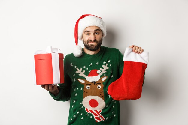 Concepto de vacaciones de Navidad e invierno. Chico confundido sosteniendo un calcetín de Navidad y una caja de regalo, encogiéndose de hombros indeciso, de pie con gorro de Papá Noel sobre fondo blanco.