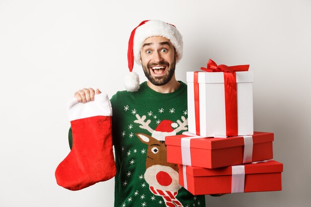 Concepto de vacaciones de Navidad e invierno. Hombre emocionado sosteniendo calcetines de Navidad y cajas de regalo, celebrando el año nuevo, de pie sobre fondo blanco.