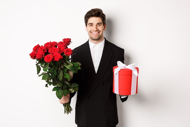 Concepto de vacaciones, relación y celebración. Novio guapo en traje negro, sosteniendo un ramo de rosas rojas y un regalo, deseando feliz navidad, de pie sobre fondo blanco.