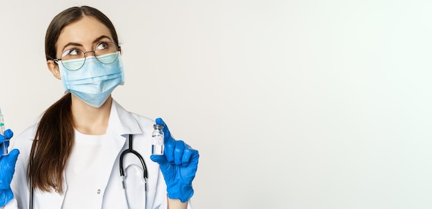 Foto gratuita concepto de vacunación y atención médica de covid19 médico joven con mascarilla médica que muestra una jeringa y una vacuna de la variante omicron del coronavirus de pie sobre fondo blanco