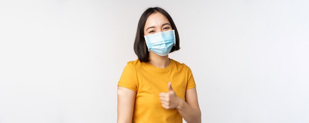 Concepto de vacunación y atención médica covid19 Retrato de una linda niña asiática con máscara médica tiene tirita en el hombro después de la vacuna contra el coronavirus de pie sobre fondo blanco