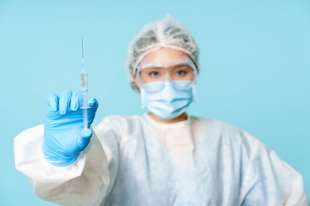 Concepto de vacunación y covid-19. Trabajador médico, médico asiático que muestra la vacuna contra el coronavirus en una jeringa, con equipo de protección personal y mascarilla, fondo azul