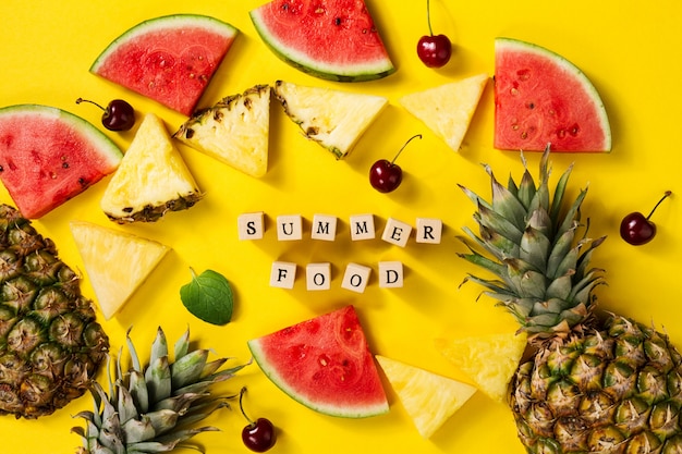 Foto gratuita concepto de verano. conceptual. sabrosas rodajas apetitosas de piña, cereza y sandía sobre fondo vibrante brillante de color amarillo con letras de madera verano. flat lay.