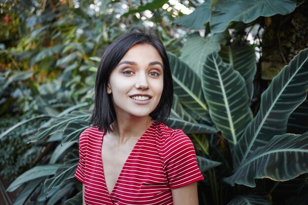 Concepto de verano. Imagen de feliz hermosa joven dama de cabello bob con amplia sonrisa encantadora descansando al aire libre, caminando en el bosque o parque, de pie entre verdes plantas exóticas y sonriendo