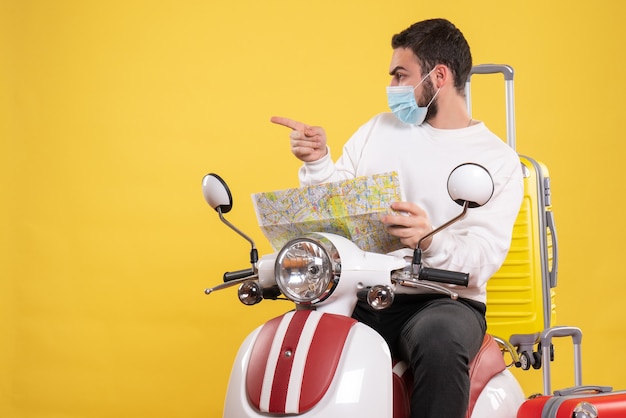 Concepto de viaje con chico curioso en máscara médica sentado en motocicleta con maleta amarilla y mostrando el mapa en amarillo