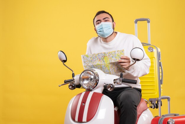 Concepto de viaje con chico curioso en máscara médica sentado en motocicleta con maleta amarilla y sosteniendo mapa en amarillo