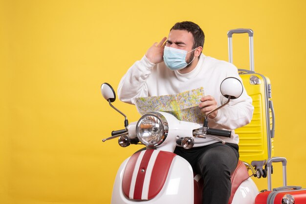 Concepto de viaje con un chico joven con máscara médica sentado en una motocicleta con una maleta amarilla y sosteniendo un mapa que sufre de dolor de cabeza