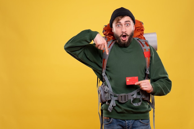 Foto gratuita concepto de viaje con chico joven sorprendido con packpack y sosteniendo la tarjeta bancaria apuntando hacia abajo en amarillo