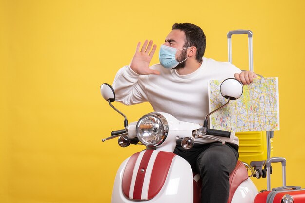 Concepto de viaje con chico nervioso en máscara médica sentado en motocicleta con maleta amarilla y mostrando mapa en amarillo