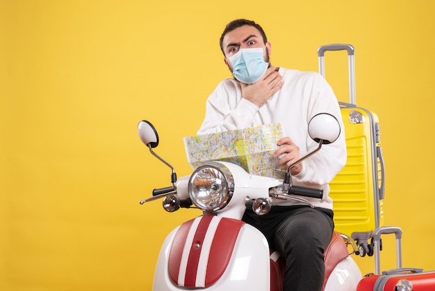 Concepto de viaje con chico nervioso en máscara médica sentado en motocicleta con maleta amarilla y sosteniendo mapa asfixiándose en amarillo