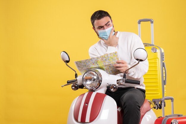 Concepto de viaje con chico pensante en máscara médica sentado en motocicleta con maleta amarilla y mostrando el mapa en amarillo