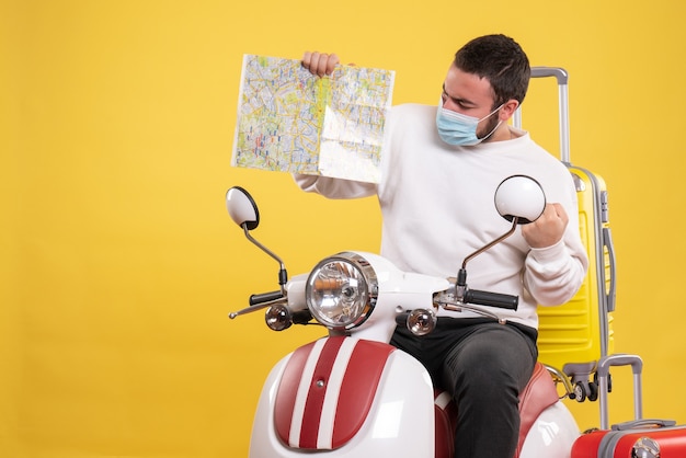 Concepto de viaje con chico seguro en máscara médica sentado en motocicleta con maleta amarilla y mostrando mapa en amarillo