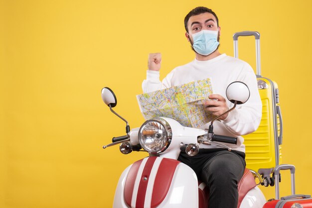 Concepto de viaje con chico seguro en máscara médica sentado en motocicleta con maleta amarilla y sosteniendo mapa en amarillo