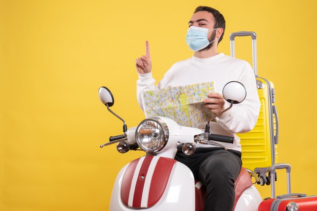 Concepto de viaje con chico seguro en máscara médica sentado en motocicleta con maleta amarilla y sosteniendo el mapa apuntando hacia arriba en amarillo