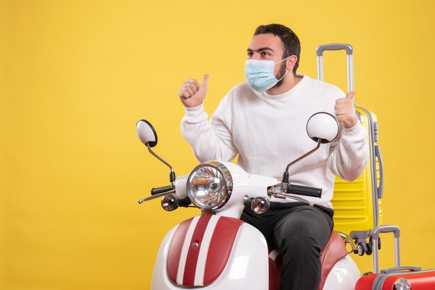 Concepto de viaje con chico sonriente en máscara médica sentado en motocicleta con maleta amarilla y haciendo gesto de ok en amarillo