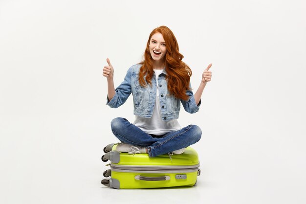 Concepto de viaje y estilo de vida. Mujer caucásica emocionada joven que se sienta en la valija del equipaje que muestra el pulgar hacia arriba. Aislado en blanco. Listo para vacaciones.