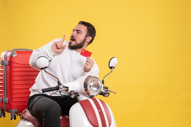 Concepto de viaje con joven hombre viajero confundido sentado en motocicleta con maleta mostrando tarjeta bancaria en amarillo