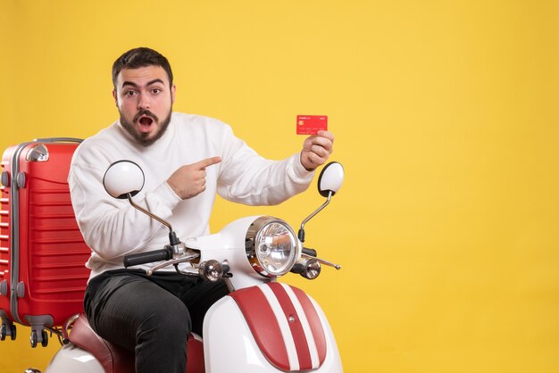 Concepto de viaje con joven viajero confundido y emocional sentado en motocicleta con maleta sosteniendo tarjeta bancaria en amarillo