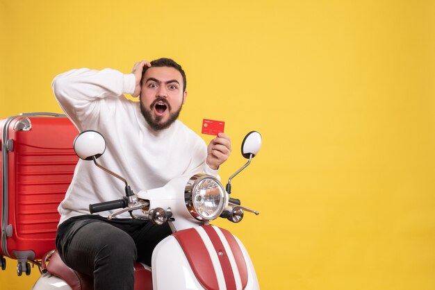 Concepto de viaje con joven viajero emocional sorprendido sentado en motocicleta con maleta sosteniendo tarjeta bancaria en amarillo